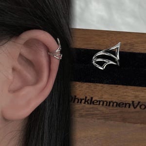 Op de afbeelding: Zilveren oormanchet met een geometrisch ontwerp. De manchet is te zien op het oor van een persoon en in een kleine houten doos.