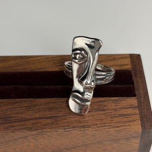 Könnte beinhalten: Ein silberfarbener Ring mit einem länglichen, abstrakten Gesichtsmotiv auf der Vorderseite. Die Metalloberfläche ist poliert und weist vertiefte Details auf, die Auge, Nase und Mund definieren. Die Ringschiene ist ein einfacher, glatter Metallring.
