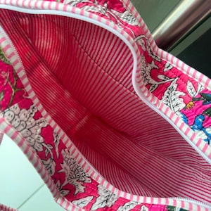 May include: A quilted tote bag featuring a vibrant pink floral block print on the exterior and a white and pink striped interior lining. The bag includes a white zipper closure along the top edge and matching striped fabric handles for carrying.