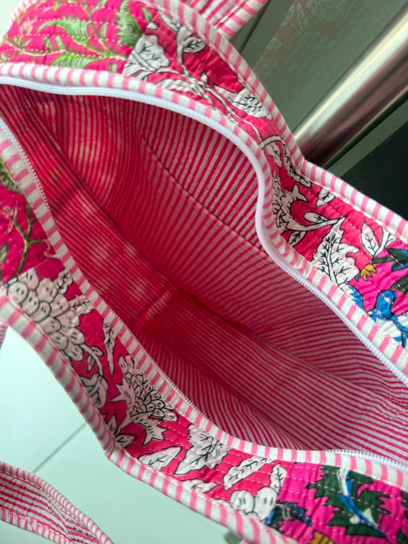 May include: A quilted tote bag featuring a vibrant pink floral block print on the exterior and a white and pink striped interior lining. The bag includes a white zipper closure along the top edge and matching striped fabric handles for carrying.