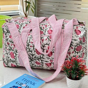 May include: A quilted tote bag with a floral pattern in pink and white. The bag has pink striped handles and a matching trim. The bag is sitting on a white surface next to a small potted plant and a book titled "Think & Grow Rich".