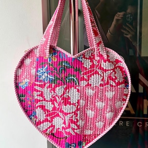 May include: A heart-shaped quilted tote bag featuring a vibrant patchwork design in shades of hot pink, light pink, and white. The fabric displays intricate floral and botanical patterns with a striped trim along the edges and two matching handles.