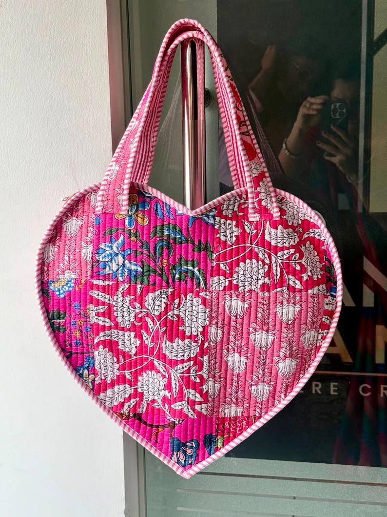 May include: A heart-shaped quilted tote bag featuring a vibrant patchwork design in shades of hot pink, light pink, and white. The fabric displays intricate floral and botanical patterns with a striped trim along the edges and two matching handles.