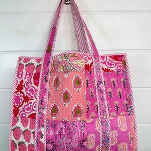 May include: A pink and white patchwork tote bag with a quilted design. The bag has a large main compartment and two shorter handles.
