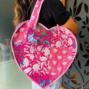 May include: A heart-shaped quilted tote bag featuring a vibrant pink patchwork design with white and blue floral block-print patterns. The bag includes two matching pink and white striped fabric handles and a striped trim along the edges.