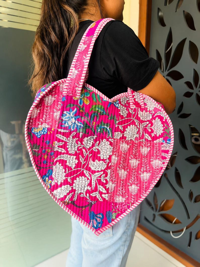May include: A heart-shaped quilted tote bag featuring a vibrant pink patchwork design with white and blue floral block-print patterns. The bag includes two matching pink and white striped fabric handles and a striped trim along the edges.