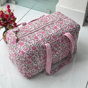 Puede incluir: Una bolsa de viaje acolchada con un estampado floral en rosa y verde sobre fondo blanco. La bolsa tiene cierre de cremallera, asas a rayas rosas y blancas y un bolsillo lateral. La bolsa mide aproximadamente 46 cm de largo, 25 cm de alto y 25 cm de ancho.