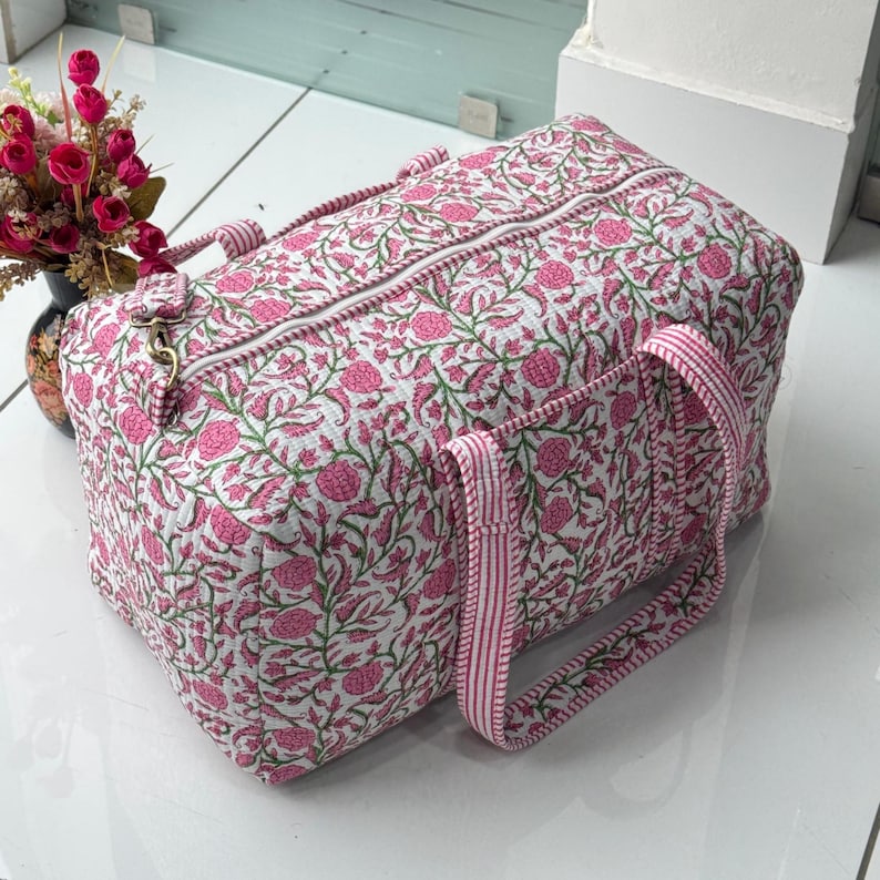 May include: A quilted travel bag with a floral pattern in pink and green on a white background. The bag has a zipper closure, pink and white striped handles, and a side pocket. The bag is rectangular and appears to be made of fabric.