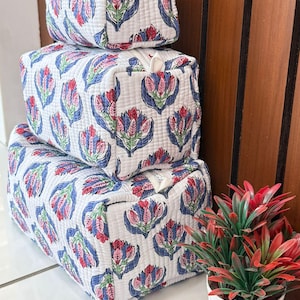 May include: Three stacked, square cosmetic bags. They feature a white background with a repeating floral pattern in red, blue, and pink. The bags have a quilted texture and zippers. A small potted plant with red and green leaves is also visible.