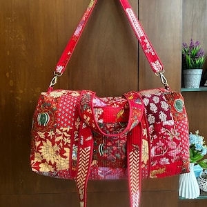 Peut inclure: Un sac de voyage rouge à motifs avec une bandoulière assortie. Le sac présente divers motifs floraux et géométriques dans des tons de rouge, or et vert. Le sac est suspendu à une porte en bois.