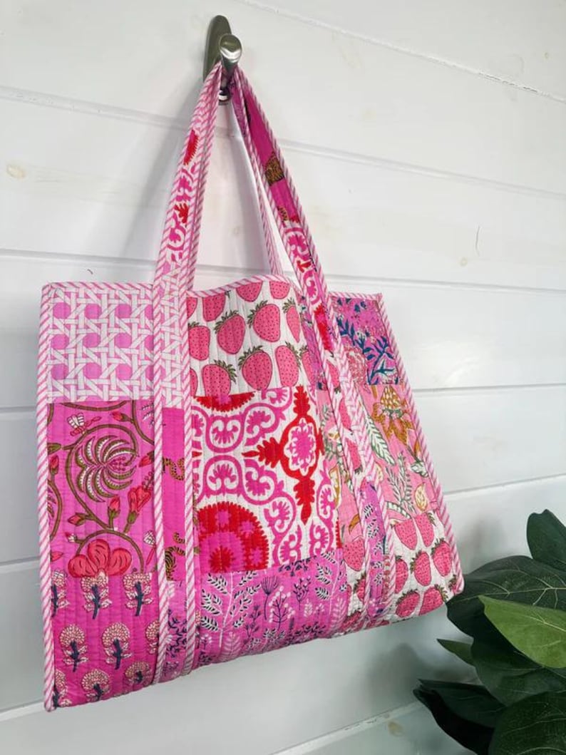 May include: A pink patchwork tote bag with white trim. The bag is made of different fabrics with floral and strawberry patterns. The bag has two handles and is hanging on a hook.