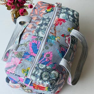May include: A quilted duffel bag featuring a patchwork design with gray, pink, blue, and yellow floral and botanical patterns. The bag has a central white zipper closure and two striped fabric handles attached to the top and side panels.
