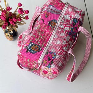 May include: A rectangular quilted duffel bag featuring a vibrant pink base with intricate floral patterns in shades of white, blue, and green. The bag includes a white zipper closure along the top and two striped pink and white fabric handles.