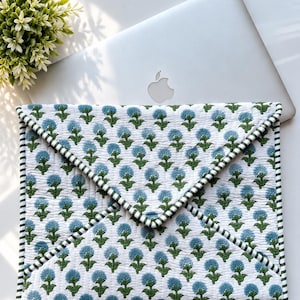 May include: A quilted laptop sleeve featuring an envelope-style flap closure with a green and white striped fabric trim. The exterior is white fabric printed with a repeating pattern of blue flower heads on green stems and leaves.