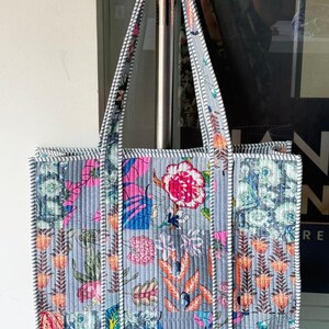 May include: A quilted tote bag with a floral pattern in shades of pink, blue, orange, and green on a gray background. The bag has two handles and is trimmed with a black and white striped border.