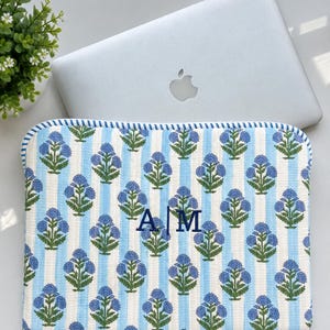 May include: A rectangular quilted laptop sleeve featuring a repeating pattern of blue floral motifs on white and light blue vertical stripes. The center of the sleeve is embroidered with the dark blue capital letters A and M separated by a vertical line.