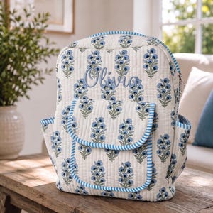 Puede incluir: Una mochila acolchada con base color crema y un patrón floral repetido en azul y verde. La bolsa incluye un bolsillo frontal con un borde a rayas azules y blancas y el nombre Olivia bordado en letra cursiva azul en la parte delantera.