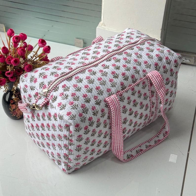 May include: A white quilted duffel bag with a repeating pink floral pattern. The bag has a zipper closure, pink and white striped handles, and a matching trim. The bag is designed for travel or carrying items.