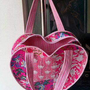 May include: A heart-shaped quilted tote bag featuring a vibrant pink floral block print pattern. The bag is constructed with a pink and white striped fabric trim along the edges and includes two matching striped fabric handles for carrying.