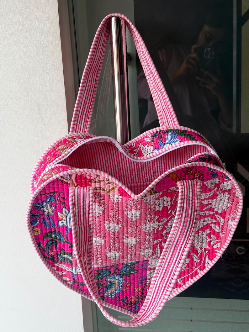 May include: A heart-shaped quilted tote bag featuring a vibrant pink floral block print pattern. The bag is constructed with a pink and white striped fabric trim along the edges and includes two matching striped fabric handles for carrying.