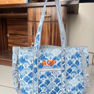 Puede incluir: Un bolso tote acolchado con un patrón de conchas azules y blancas con bordes laterales e inferiores con volantes. El bolso tiene dos correas largas para el hombro y un bolsillo frontal con cremallera con las letras AQP bordadas en naranja.