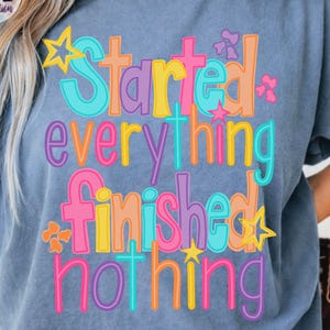 Może przedstawiać: Niebieska koszulka z napisem "Started everything finished nothing" w kolorowych literach. Projekt zawiera gwiazdy i kwiatowe akcenty, tworząc wesoły i przyciągający wzrok wygląd.