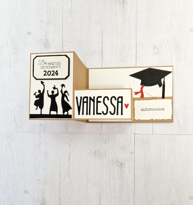 May include: A graduation card with a pop-up design. The card is brown with white accents. The front of the card has a black silhouette of three people throwing their caps in the air. The text on the card reads "Du hast es geschafft! 2024". The inside of the card has a pop-up graduation cap and the text "Vanessa" with a red heart. The card also has a small tag that says "Gl&uuml;ckwunsch".