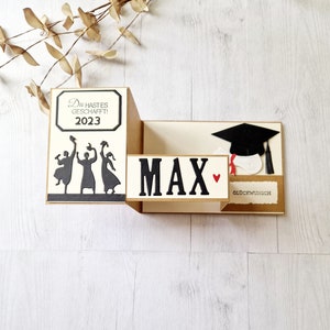 May include: A graduation card with a pop-up design. The card features a black silhouette of three people throwing their caps in the air, a black graduation cap, and the text "Du hast es geschafft! 2023" and "MAX" with a red heart.
