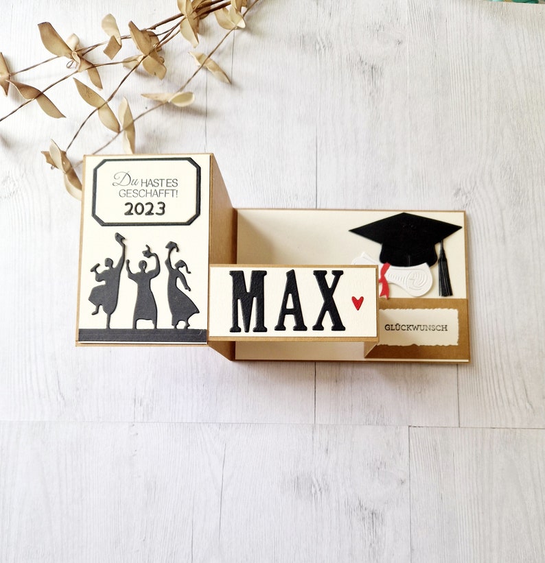 May include: A graduation card with a pop-up design. The card features a black silhouette of three people throwing their caps in the air, a black graduation cap, and the text "Du hast es geschafft! 2023" and "MAX" with a red heart.