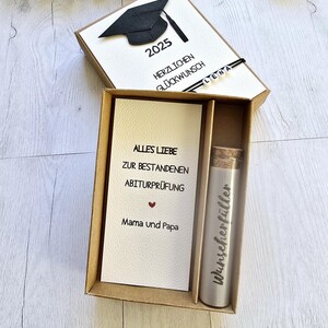 May include: A brown box with a white card inside. The card has a black graduation cap and the text "2025" and "Herzlichen Gl&uuml;ckwunsch". The box also contains a small silver tube with the text "Wunschersch&ouml;pfer" on it. The card inside the box has the text "Alles Liebe zur bestandenen Abiturpr&uuml;fung Mama und Papa".