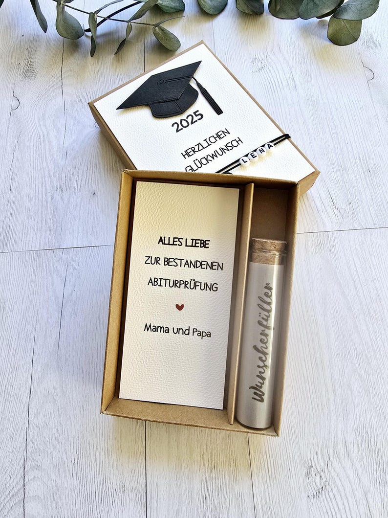 May include: A brown box with a white card inside. The card has a black graduation cap and the text "2025" and "Herzlichen Gl&uuml;ckwunsch". The box also contains a small silver tube with the text "Wunschersch&ouml;pfer" on it. The card inside the box has the text "Alles Liebe zur bestandenen Abiturpr&uuml;fung Mama und Papa".