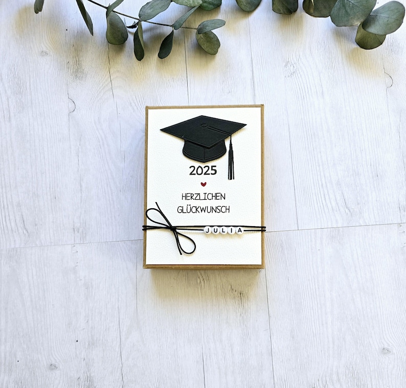 May include: A white card with a black graduation cap, the year 2025, a red heart, and the text "Herzlichen Gl&uuml;ckwunsch" and "JULIA".