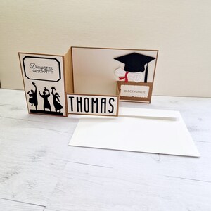 May include: A brown card with a pop-up graduation cap and diploma. The card says "Du hast es geschafft!" and "Gl&uuml;ckwunsch" and has a silhouette of people throwing their caps in the air. The card is open to reveal the name "Thomas" on the inside.
