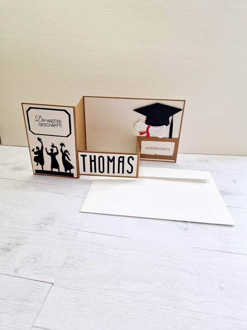 May include: A brown card with a pop-up graduation cap and diploma. The card says "Du hast es geschafft!" and "Gl&uuml;ckwunsch" and has a silhouette of people throwing their caps in the air. The card is open to reveal the name "Thomas" on the inside.