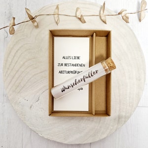 May include: A small glass tube with a cork stopper, labeled "Wunscheref&uuml;ller" and "-pa". The tube is in a brown cardboard box with a white card that reads "Alles Liebe zur bestandenen Abiturpr&uuml;fung".