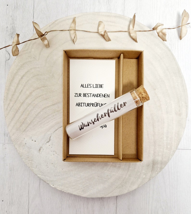 May include: A small glass tube with a cork stopper, labeled "Wunscheref&uuml;ller" and "-pa". The tube is in a brown cardboard box with a white card that reads "Alles Liebe zur bestandenen Abiturpr&uuml;fung".