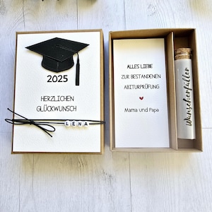 May include: A brown box with a white card inside. The card has a black graduation cap with the year 2025 printed below it. The text "Herzlichen Gl&uuml;ckwunsch" is printed below the cap. The text "Alles Liebe zur bestandenen Abiturpr&uuml;fung" is printed on the card in the other box. The text "Mama und Papa" is printed below the text. A small glass vial with a cork stopper is in the other box. The text "Wunsch erf&uuml;ller" is printed on the vial.