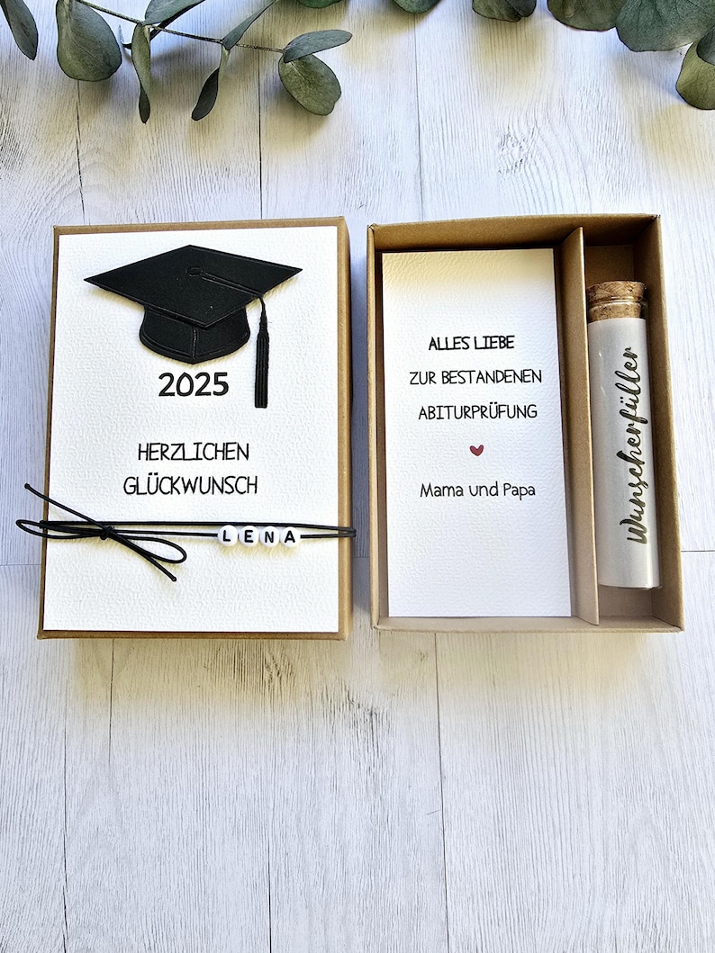May include: A brown box with a white card inside. The card has a black graduation cap with the year 2025 printed below it. The text "Herzlichen Gl&uuml;ckwunsch" is printed below the cap. The text "Alles Liebe zur bestandenen Abiturpr&uuml;fung" is printed on the card in the other box. The text "Mama und Papa" is printed below the text. A small glass vial with a cork stopper is in the other box. The text "Wunsch erf&uuml;ller" is printed on the vial.