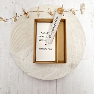 May include: A small glass vial with a cork stopper, labeled "Wunscherrf&uuml;ller" in black lettering. The vial is sitting in a brown cardboard box with a white card that reads "Alles Liebe zur bestandenen Abiturpr&uuml;fung Mama und Papa" with a small red heart.