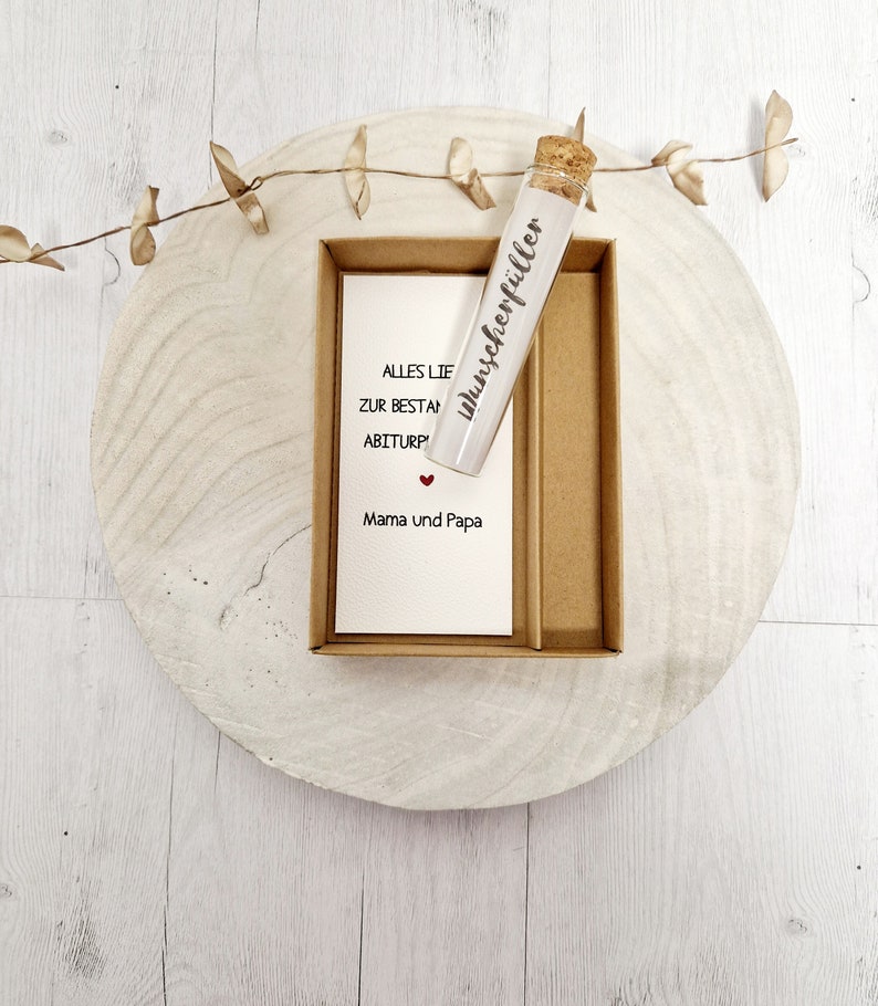 May include: A small glass vial with a cork stopper, labeled "Wunscherrf&uuml;ller" in black lettering. The vial is sitting in a brown cardboard box with a white card that reads "Alles Liebe zur bestandenen Abiturpr&uuml;fung Mama und Papa" with a small red heart.