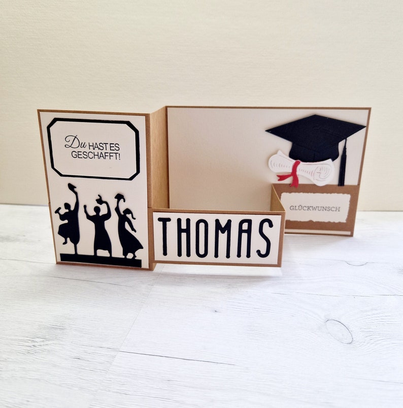 May include: A graduation card with a brown background and a white interior. The card features a silhouette of three people throwing their caps in the air, the text "Du hast es geschafft!" and the name "Thomas". The inside of the card has a black graduation cap with a diploma and the text "Gl&uuml;ckwunsch".