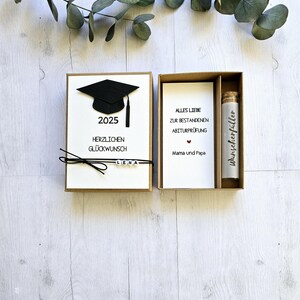 May include: A brown box with a white card inside. The card has a black graduation cap, the year 2025, the text "Herzlichen Gl&uuml;ckwunsch" and the name "Lena". The box also contains a smaller white card with the text "Alles Liebe zur bestandenen Abiturpr&uuml;fung Mama und Papa" and a small glass bottle with a cork stopper and the text "Wunschersf&uuml;ller".