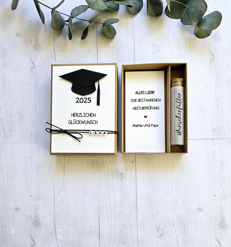 May include: A brown box with a white card inside. The card has a black graduation cap, the year 2025, the text "Herzlichen Gl&uuml;ckwunsch" and the name "Lena". The box also contains a smaller white card with the text "Alles Liebe zur bestandenen Abiturpr&uuml;fung Mama und Papa" and a small glass bottle with a cork stopper and the text "Wunschersf&uuml;ller".