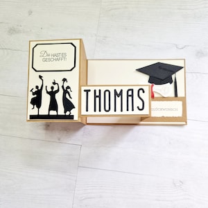 May include: A graduation card with a pop-up design featuring three figures throwing their caps in the air. The card is decorated with a black graduation cap, a diploma, and the text "Thomas" and "Gl&uuml;ckwunsch".
