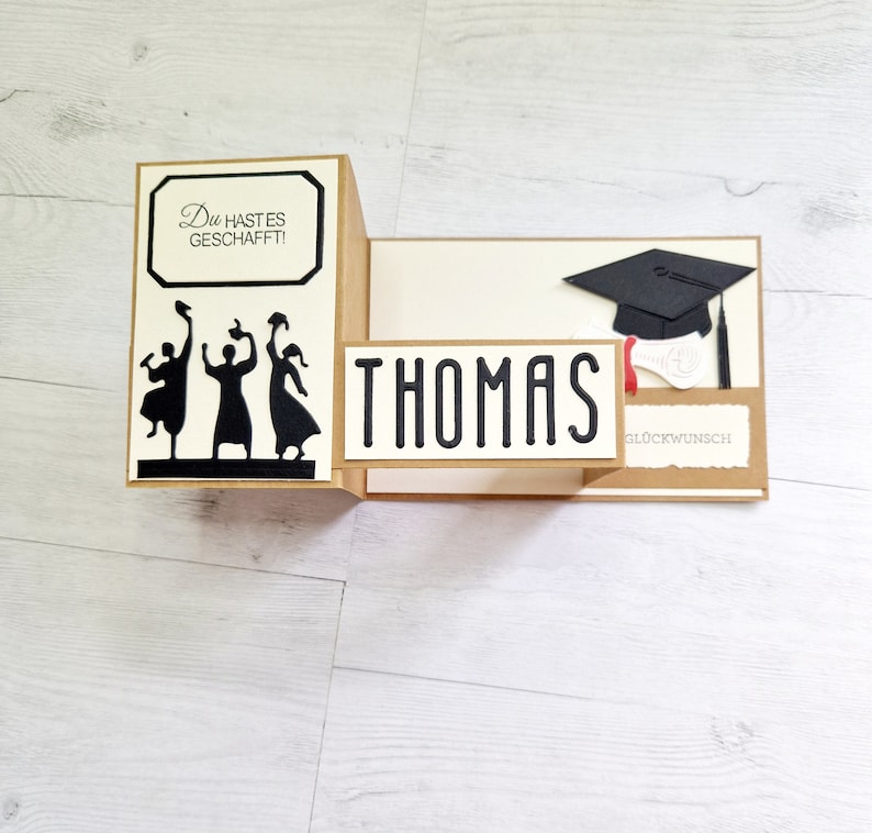 May include: A graduation card with a pop-up design featuring three figures throwing their caps in the air. The card is decorated with a black graduation cap, a diploma, and the text "Thomas" and "Gl&uuml;ckwunsch".