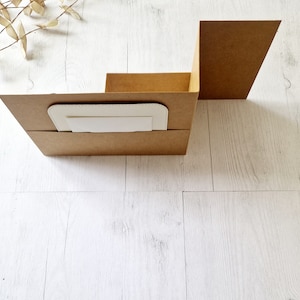 May include: A brown cardboard box with a white insert. The box is folded into a vertical shape, with the top flap open. The box is sitting on a white wooden surface.