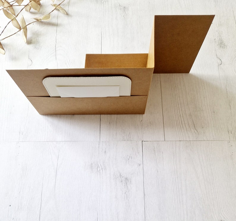 May include: A brown cardboard box with a white insert. The box is folded into a vertical shape, with the top flap open. The box is sitting on a white wooden surface.