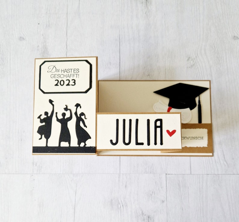 May include: A graduation card featuring a black silhouette of three people throwing their caps in the air. The card reads "Du hast es geschafft! 2023" and "Julia" with a red heart. The card also includes a black graduation cap with a tassel.