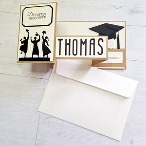 May include: A graduation card with a pop-up design featuring three figures throwing graduation caps in the air. The card is decorated with a black graduation cap and the name "THOMAS" in black lettering. The card also includes the text "Du hast es geschafft!" and "Wunsch".