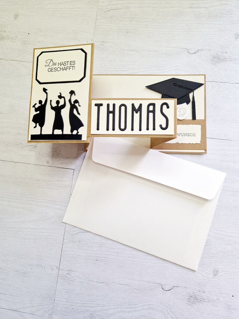 May include: A graduation card with a pop-up design featuring three figures throwing graduation caps in the air. The card is decorated with a black graduation cap and the name "THOMAS" in black lettering. The card also includes the text "Du hast es geschafft!" and "Wunsch".